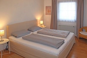 Ein Schlafzimmer mit einem Bett, einem Nachttisch mit Lampe, einem Sessel und einem Fenster mit Vorhängen.
