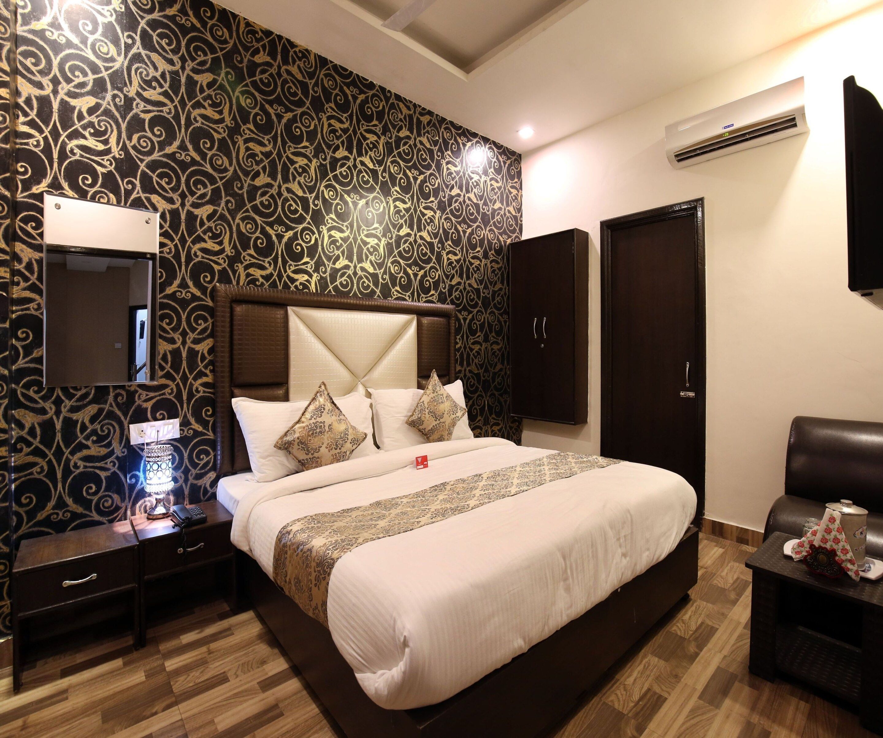 A hotel room with a large bed, a nightstand, a lamp, a wardrobe, and an air conditioning unit.