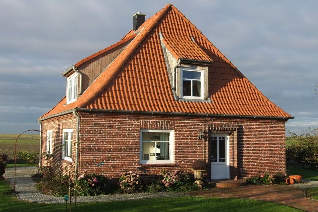 Une maison à deux étages, construite en briques, avec un toit recouvert de tuiles rouges, une porte d’entrée et un petit porche agrémenté d’un pot de fleurs.