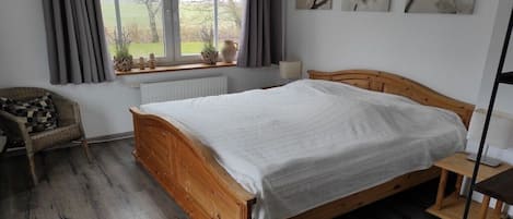 A bedroom with a wooden bed, a window with curtains, a radiator, a chair, and wall art.