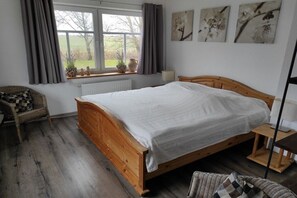 A bedroom with a wooden bed, a window with curtains, a radiator, a chair, and wall art. - Lüdde Hus Mitteldiek (Nordstrand)