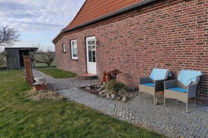 A brick house with a red roof, a small shed, and two wicker chairs with cushions. - Lüdde Hus Mitteldiek (Nordstrand)