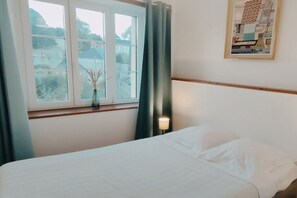 Ein Hotelzimmer mit Bett, einem Fenster mit Vorhängen, einer Vase mit Trockenpflanzen und einem gerahmten Bild an der Wand.