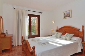 A bedroom with a wooden bed, a nightstand, a dresser, a window with curtains, and a wall-mounted lamp.