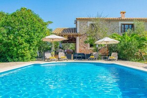 A swimming pool with lounge chairs and umbrellas, surrounded by greenery and a stone building.
