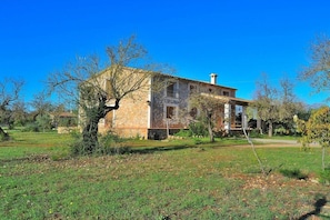 A two-story stone building with a balcony, surrounded by a grassy area and trees.