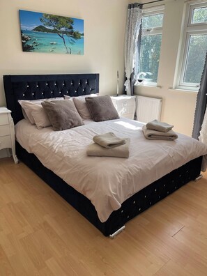 A neatly made bed with a dark headboard, a white bedside table, and a painting of a coastal scene on the wall. - The Cosy Nest - Overseal (Swadlincote)