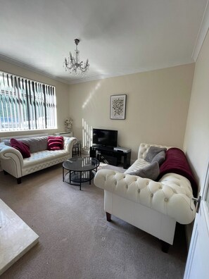 A living room with a white sofa, a smaller white armchair, a glass coffee table, a TV stand with a flat-screen TV, and a chandelier. - The Cosy Nest - Overseal (Swadlincote)