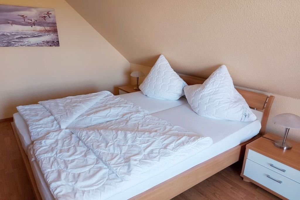 Cama bem arrumada com edredom branco e almofadas, luminária de cabeceira e uma cômoda pequena.