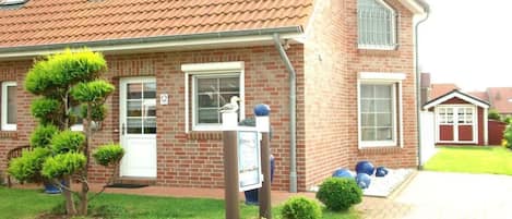 Une maison en briques avec un toit en tuiles, un petit jardin avec un arbre et des arbustes, et une boîte aux lettres.