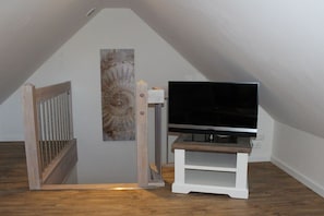 Ein Zimmer mit schrägen Wänden, einer Holztreppe, einem Flachbildfernseher auf einem weißen Standfuß und einem an der Wand befestigten Kunstwerk.