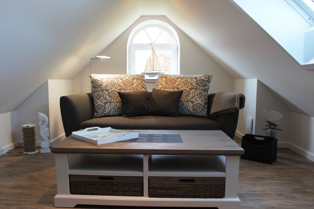 A cozy loft bedroom with a sloped ceiling, a large window, a sofa with patterned cushions, a wooden coffee table, and storage baskets.