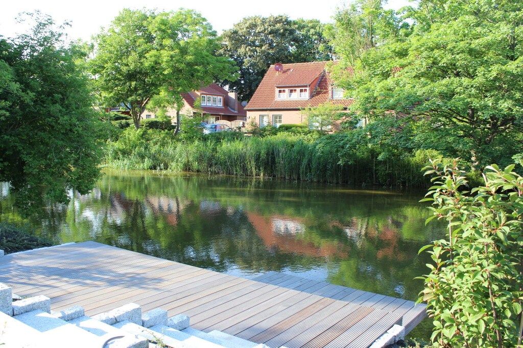 Une jetée en bois s’étendant sur une étendue d’eau calme, où se reflètent les arbres et les maisons environnants.