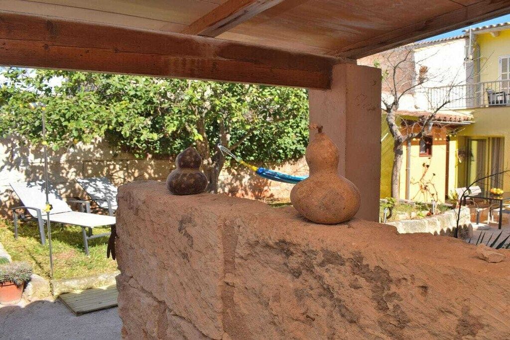 Un espacio al aire libre rústico con un muro de piedra, una pérgola de madera y un edificio amarillo al fondo.