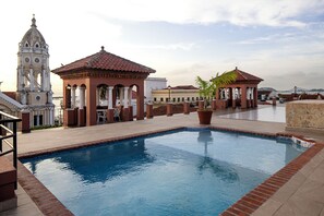 Une piscine dont le bord est revêtu de carreaux, entourée d’un chemin pavé en briques et jouxtant un bâtiment dont le toit est recouvert de tuiles rouges.