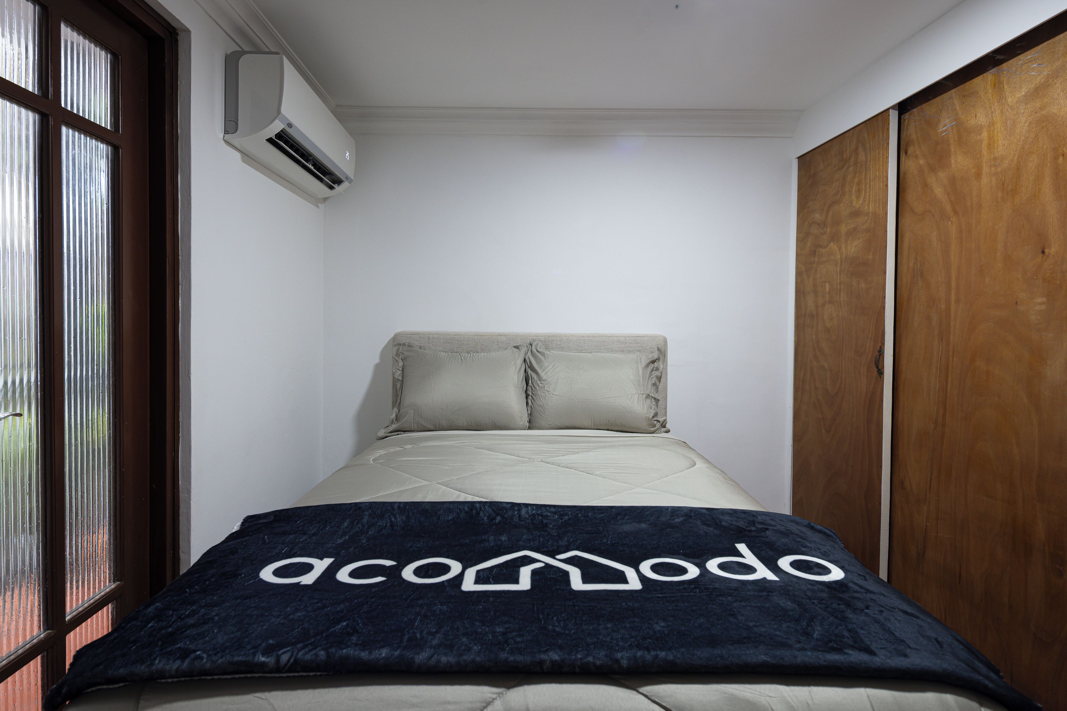 Une petite pièce étroite, avec un lit, une fenêtre et un appareil de climatisation fixé au mur.