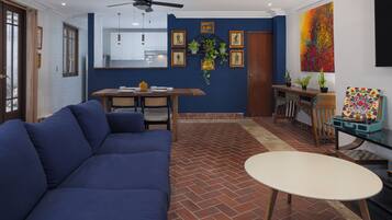 A living room with a blue sofa, a dining table with chairs, a small round table, and a ceiling fan.