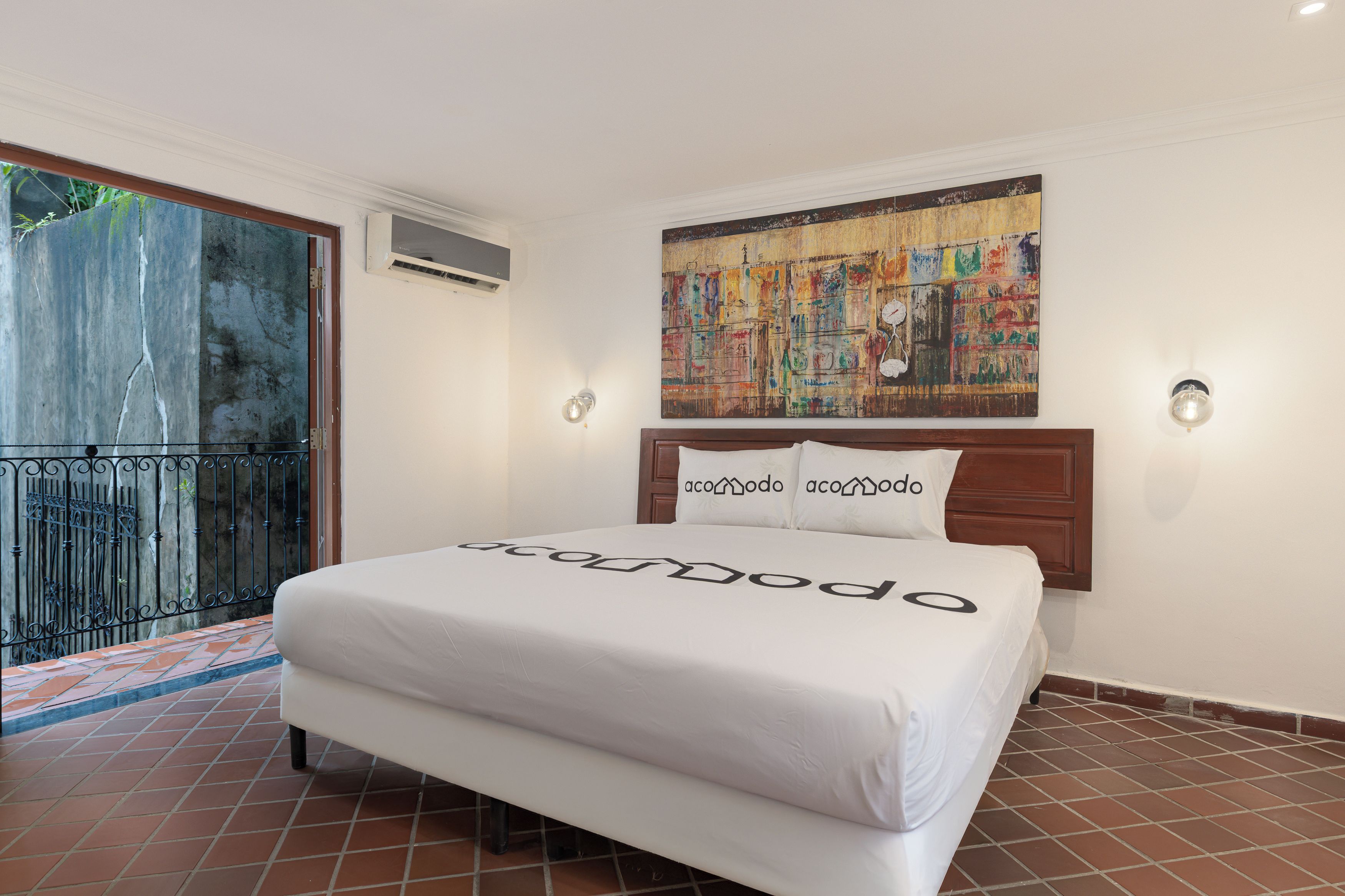 A hotel room with a bed, a painting above the bed, and a balcony with a railing.