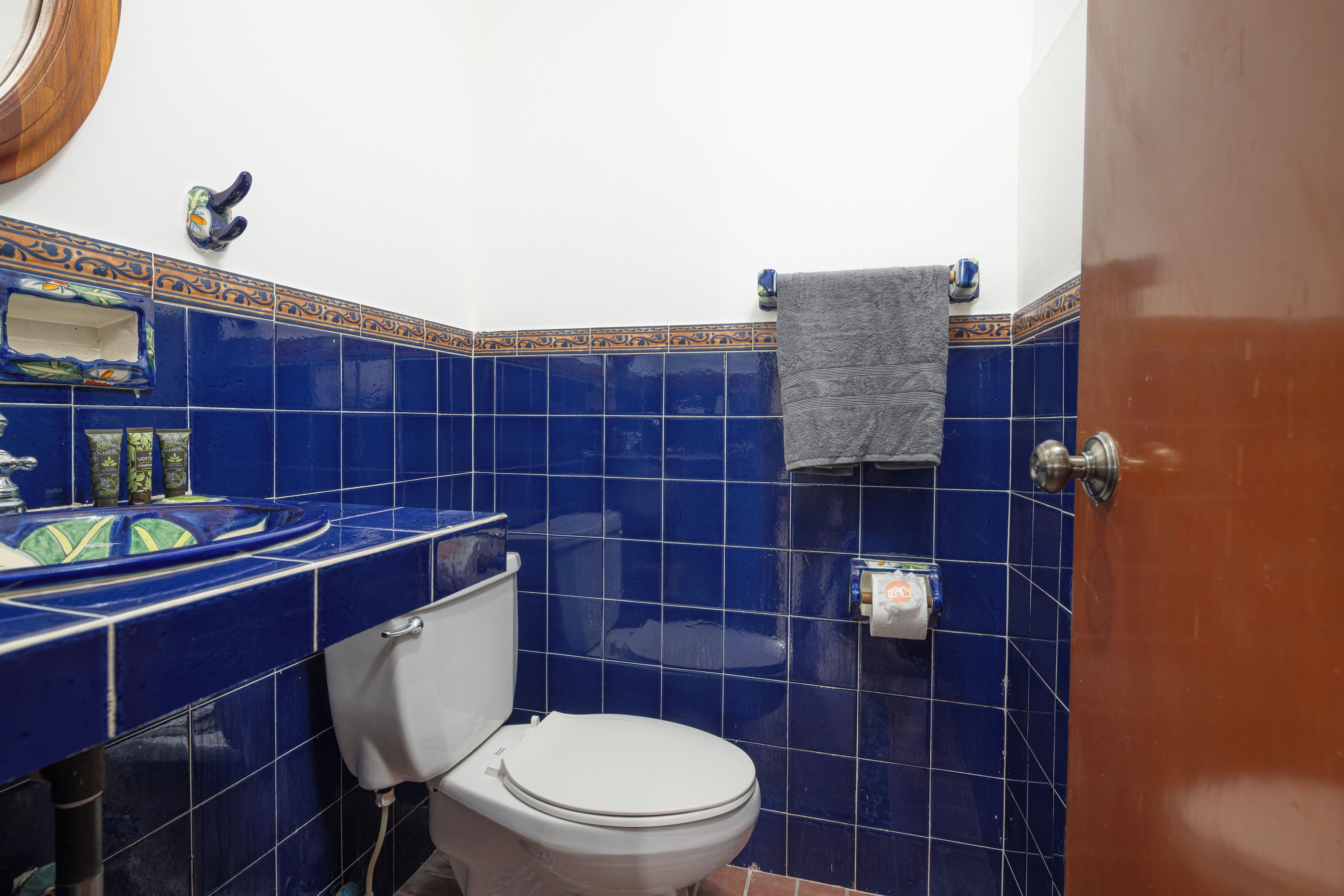 A bathroom with a white toilet, blue tiles, a sink with a decorative mirror, and a towel hanging on the wall.
