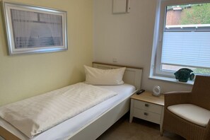 A small, single-bed room with a bedside table, a chair, and a window with blinds. - Holiday apartment with 2 bedrooms (Krummhörn-Greetsiel)
