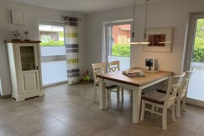 A dining area with a wooden table, white chairs, a cabinet, and a window with curtains. - Holiday apartment with 2 bedrooms (Krummhörn-Greetsiel)