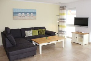 A living room with a sectional sofa, a coffee table, a TV, and a painting of beach chairs. - Holiday apartment with 2 bedrooms (Krummhörn-Greetsiel)
