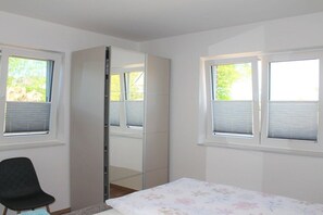 Ein Schlafzimmer mit einem Bett, einem Sessel, einem Kleiderschrank und zwei Fenstern mit Jalousien.