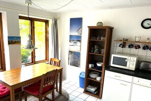 Un coin repas avec une table et des chaises en bois, une poubelle bleue, un four à micro-ondes blanc et une étagère avec des tasses.