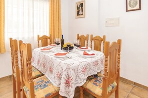 Ein Esszimmer mit einem für sechs Personen gedeckten Tisch, Holzsesseln, einer Tischdecke mit Blumenmuster und einem Fenster mit Vorhängen.