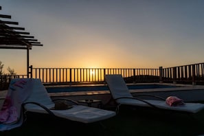 Une vue sur le coucher de soleil depuis un espace aménagé au bord de la piscine, avec des chaises longues et une structure en forme de pergola.