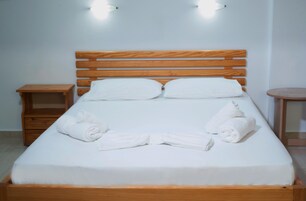 Una cama bien hecha con sábanas blancas y dos almohadas, una mesita de noche de madera con un cajón, y una pequeña mesa redonda con una lámpara.