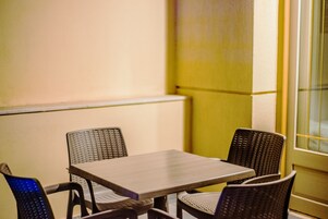 A corner room with a table and four wicker chairs, a window with blinds, and a plain wall.