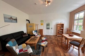 Ein Wohnzimmer mit einer Couch, Couchtisch und Sesseln, einem Holzespeisetisch mit Stühlen, einem Bücherregal und einer Tür mit Milchglasfenster.
