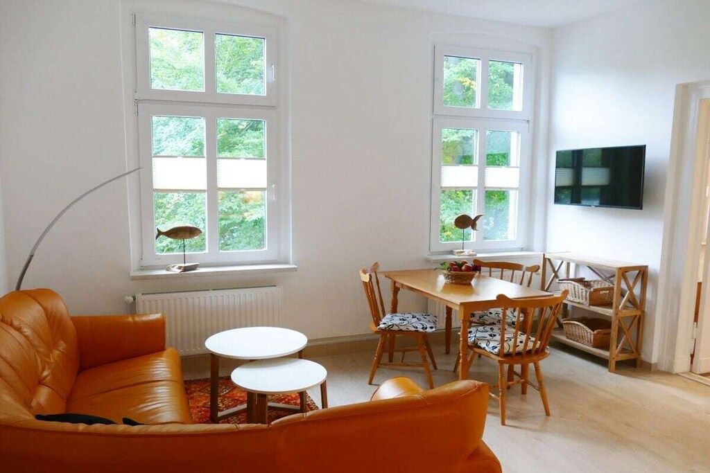 Una sala de estar con un sofá, una mesa de comedor con sillas, un televisor montado en la pared y ventanas con vistas a zonas verdes.