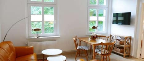 Una sala de estar con un sofá, una mesa de comedor con sillas, un televisor montado en la pared y ventanas con vistas a zonas verdes.