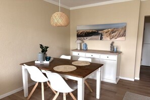 A dining area with a wooden table, white chairs, a woven pendant light, a painting of a beach scene, and a white cabinet with a lamp and decorative items. - Holiday apartment Meerweh (Bojendorf\/Fehmarn)