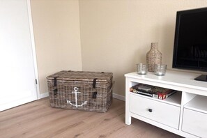 A room with a wicker storage box, a white TV stand with a flat-screen TV, and a few glass items on top. - Holiday apartment Meerweh (Bojendorf\/Fehmarn)
