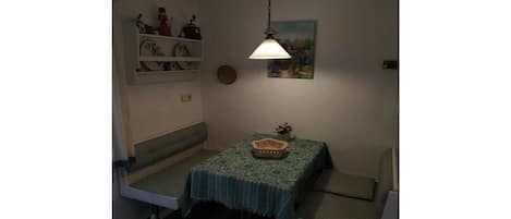 A small room with a table covered by a patterned tablecloth, a bench, a hanging light fixture, and a wall-mounted shelf with decorative items.