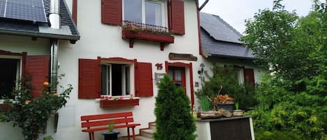 Un edificio de dos pisos con contraventanas rojas, un banco y plantas en macetas.