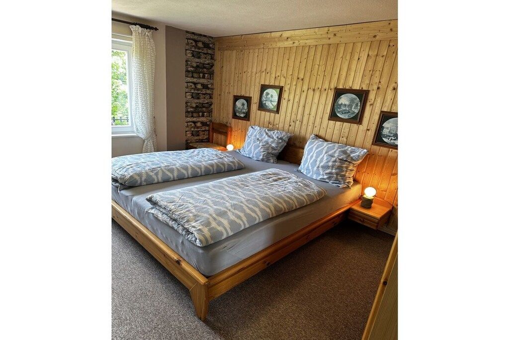 Een tweepersoonsbed met houten frame en grijze beddengoed, een stenen muur, houten wandbekleding en ingelijste foto's aan de muur.