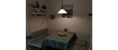 A small room with a table covered by a patterned tablecloth, a bench, a hanging light fixture, and a wall-mounted shelf with decorative items.