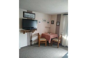 Une pièce avec un téléviseur posé sur un meuble en bois, une petite table recouverte d’une nappe rose, deux chaises en bois et une lampe.