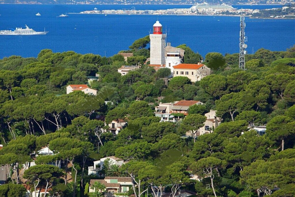 A coastal town with a prominent lighthouse, dense greenery, and a cruise ship in the background.