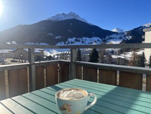 A balcony with a table and a cup of coffee, overlooking a snowy mountain and a town. - Satin Home Appartement/Fewo, Bad, WC Studio - 3 Betten by Interhome (Savognin)