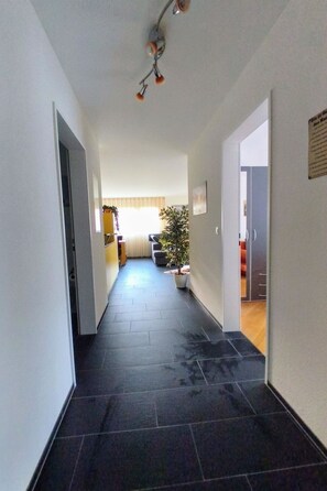 Corredor com piso ladrilhado que dá para uma sala de estar com sofá e planta.