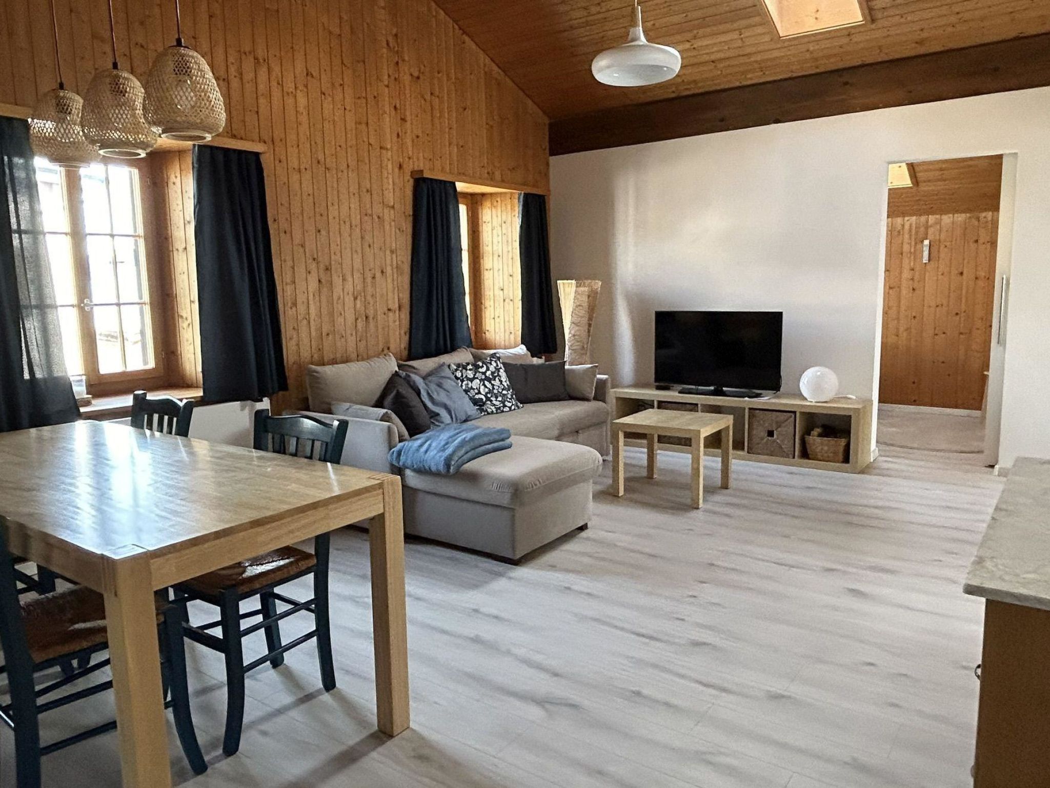 A spacious living area with a wooden floor, a sofa set, a TV, and a dining table.