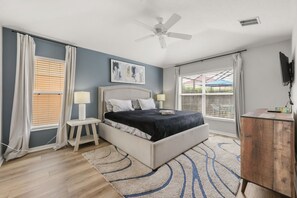 Ein Schlafzimmer mit einem Bett, Nachttischen, einem Deckenventilator, einem Fenster mit Jalousien und einem an der Wand montierten Fernseher.