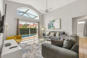 Ein geräumiges Wohnzimmer mit einem großen grauen Sofa, einem gelben Sessel, einem Glastisch und einem Deckenventilator.