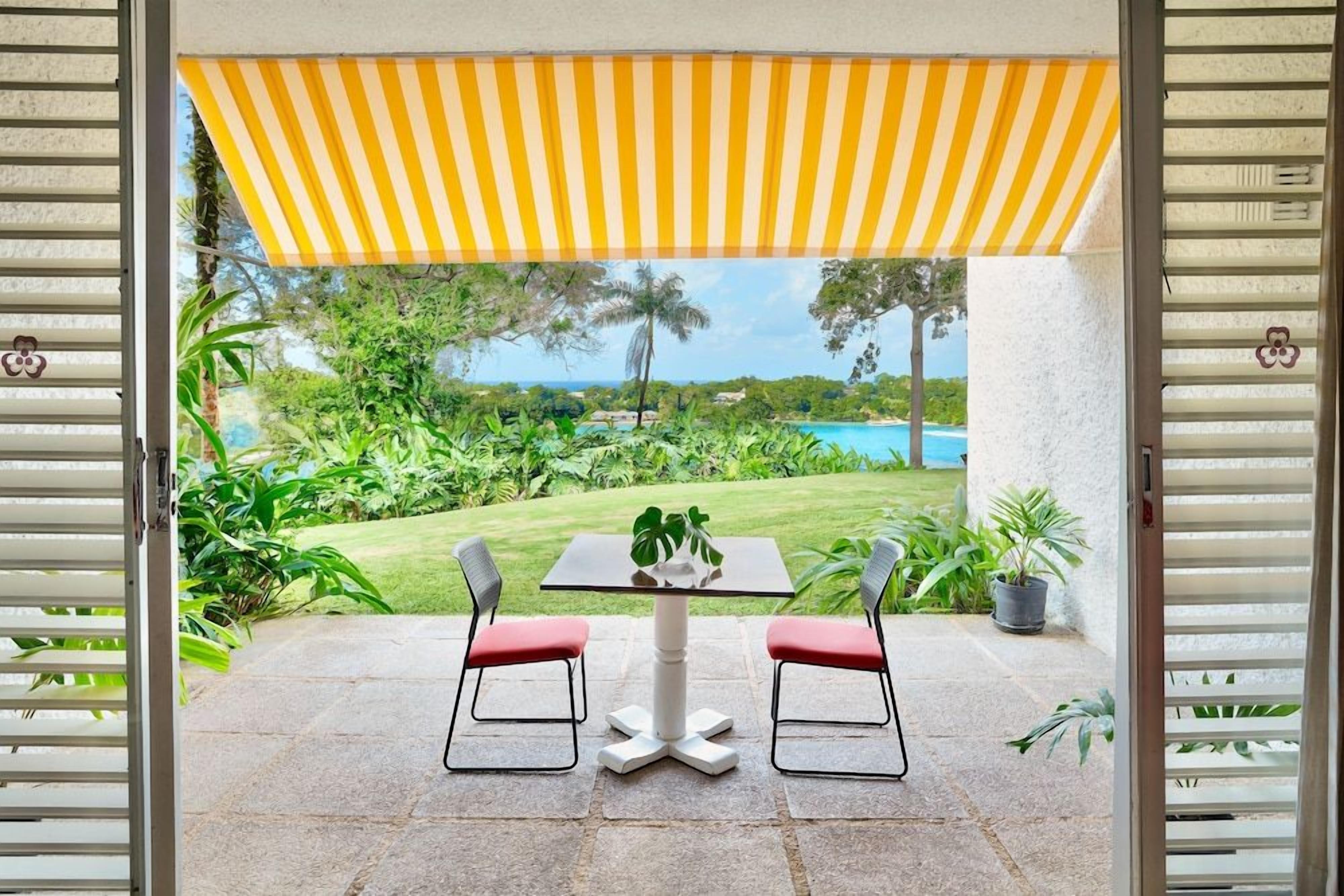 An outdoor patio with a table and two chairs, a yellow striped awning, and a view of a lush garden and a pool.