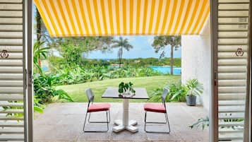 An outdoor patio with a table and two chairs, a yellow striped awning, and a view of a lush garden and a pool.